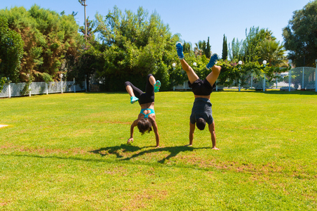 Young slim athletic couple in sportswear doing handstands on the grass, walking upside-down using...