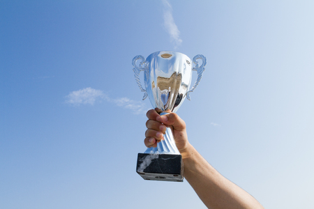 Male athlete holding up a trophy cup.  Competition winner athlete displaying champion trophy cup on blue sky background. Sports, achievement, success concept.の写真素材
