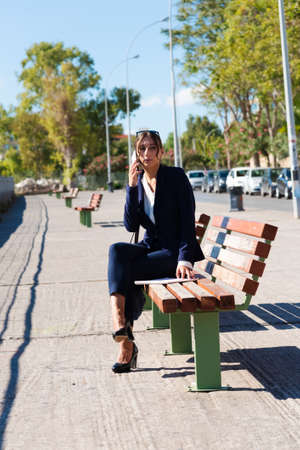 Elegant bussiness woman in marine blue suit sitting on urban street bench using mobile cell phoneの写真素材