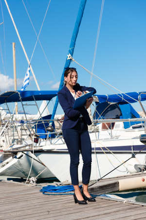 Female yachting dealer in marine blue suit holding notepad, standing outside a luxury sailboat, looking at wrist watch. Heraklion, Crete, Greeceの写真素材