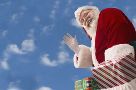 Santa Claus with bag of presents waves against a background of Cumulus clouds in an azure blue skyの写真素材