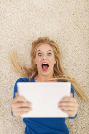 Surprise young woman lying on carpet and using digital tabletの写真素材