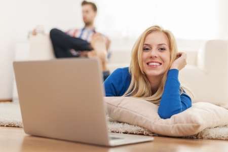 Beautiful blonde woman lying down on carpet at home and using laptopの写真素材