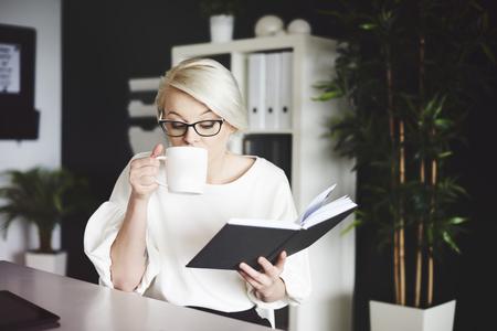 Woman reading book and drinking coffee at her deskの写真素材