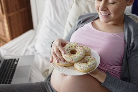 Pregnant woman eating colorful donuts in bedの写真素材