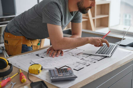 Leaning carpenter among the blueprint working at a laptop
