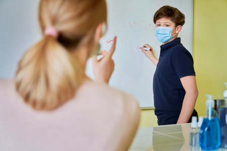 Schoolboy while answering at the blackboard wearing a protective maskの写真素材