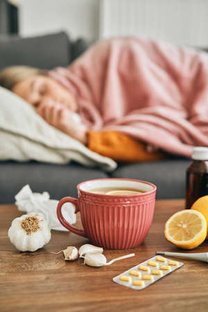Vertical image of hot tea, medicines and ill woman in the background lying on sofaの写真素材
