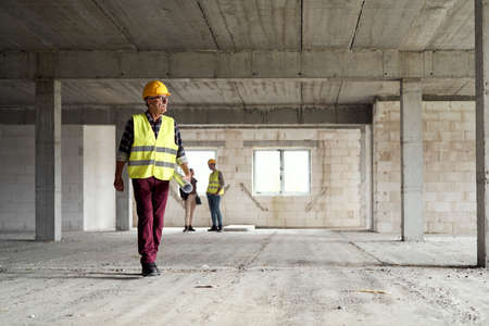 Wide image of caucasian senior engineer walking and holding plans on construction siteの写真素材