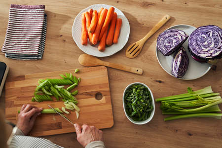 Top view of unrecognizable caucasian senior woman cutting celery while cooking in domestic kitchen