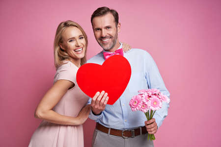 Caucasian ethnicity couple on pink background holding flowers and wooden heartの写真素材
