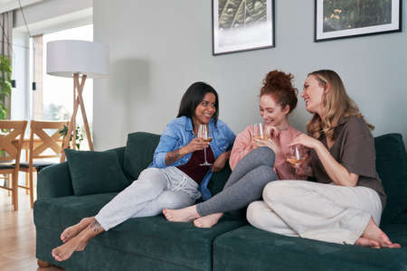 Three female caucasian friends chatting and drinking wine at homeの写真素材