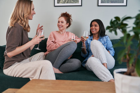 Three female caucasian friends chatting and drinking wine at homeの写真素材