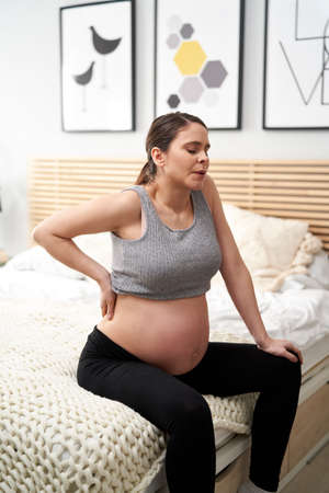 Caucasian woman in advanced pregnancy sitting on bed and feeling strong backache