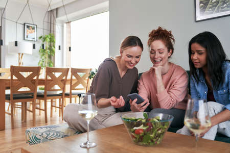 Three caucasian friends smiling while browsing smart phone on the sofaの写真素材
