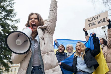 Caucasian woman screaming through megaphone in front and group of young people manifesting against Ukrainian war in the backgroundの写真素材