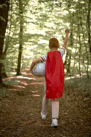 Rear view of caucasian boy wearing superhero costume walking with globe in the forestの写真素材