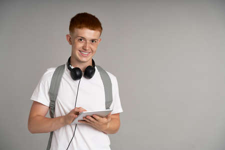 Ginger male student using digital tablet at gray background