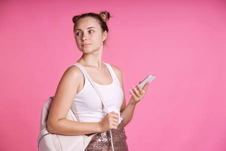 Teenage female student using mobile phone on pink background and looking awayの写真素材