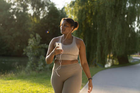 Plus sized African American woman using mobile phone and earphones during the walking at the park in...