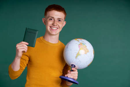 Smiling caucasian teenage boy on green background holding globe and a passport
