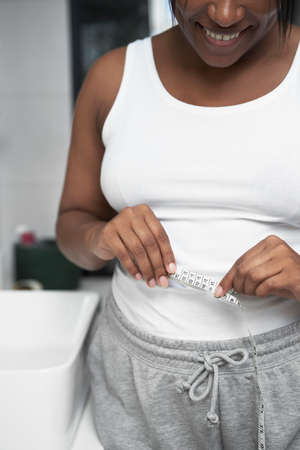 Close up of unrecognizable African-American woman standing in the bathroom and taking measures on her waistの写真素材