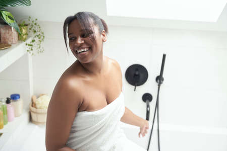 African-American woman sitting on bath wrapped in white towel and smiling
