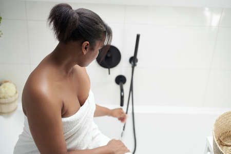 African-American woman sitting on bath wrapped in white towel and smiling
