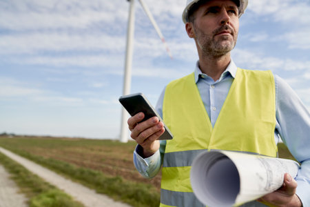 Caucasian engineer holding mobile phone and paper project while standing on wind turbine field