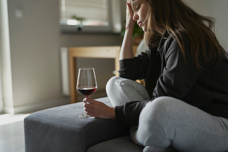Caucasian woman with alcoholism sitting at the sofa and holding glass of wineの写真素材