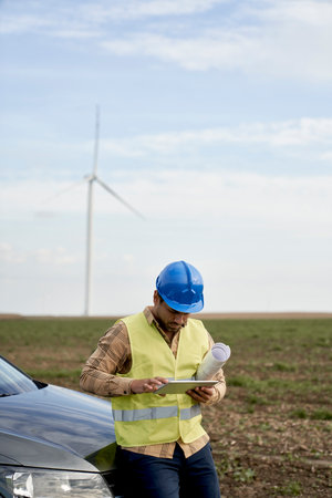 Latin male engineer holding digital tablet and standing on wind turbine field with paper projectの写真素材