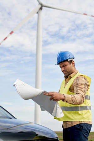 Latin male engineer standing on wind turbine field and checking paper project