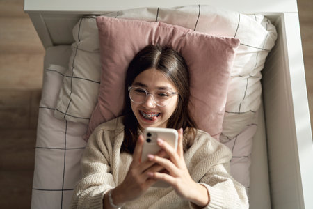 Top view of caucasian teenage girl taking selfie with phone while lying on bed on her backの写真素材