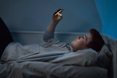 Cheerful caucasian teenage boy watching mobile phone while lying in bed at night in his room
