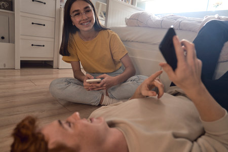 Two caucasian teenage friends having fun while using mobile phones in bedroomの写真素材