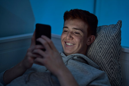 Cheerful caucasian teenage boy using mobile phone while lying in bed at night in his roomの写真素材