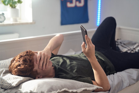 Shocked caucasian teenage boy covering eyes while browsing phone on bedの写真素材