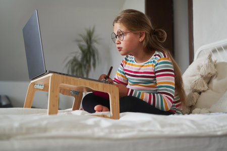 Caucasian elementary age girl studying on laptop while sitting on the bed