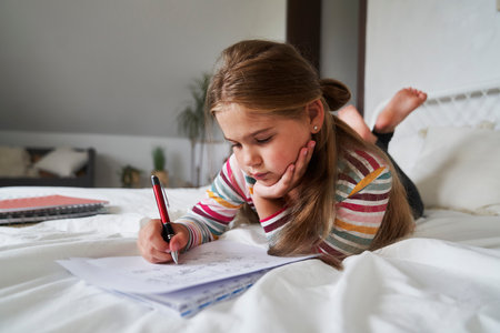 Elementary age girl studying while lying on the bedの写真素材