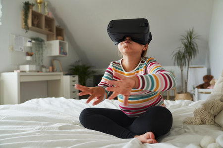 Elementary age girl sitting on bed and using virtual reality glassesの写真素材