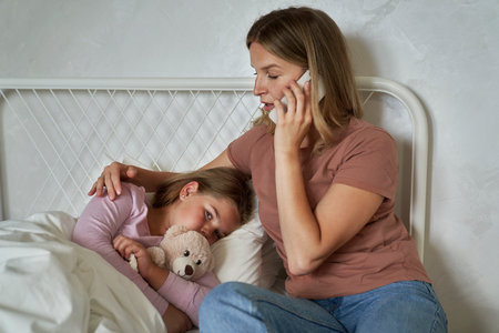 Mother calling a doctor while her daughter lying ill in bedの写真素材
