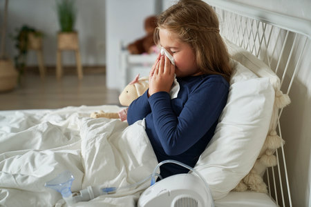 Ill elementary age girl blowing nose while sitting on bedの写真素材