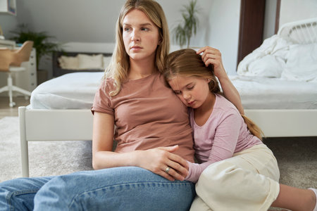 Worries mother embracing her sad elementary age girl sitting on floorの写真素材