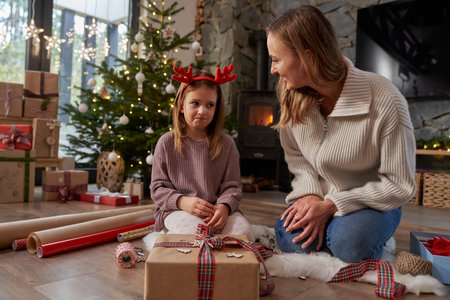 Unhappy girl and mother wrapping Christmas gifts on the floorの写真素材