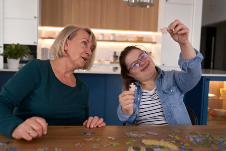 Joyful down syndrome woman and her mother assembling puzzles at homeの写真素材