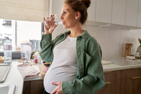 Pregnant woman drinking water while standing in the kitchenの写真素材