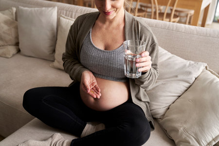 Caucasian pregnant woman sitting on sofa and taking nutritional supplementsの写真素材