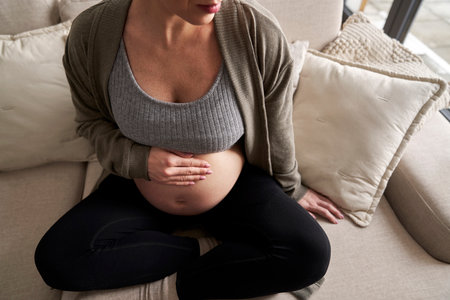 Unrecognizable caucasian pregnant woman sitting on sofa and touching her abdomenの写真素材