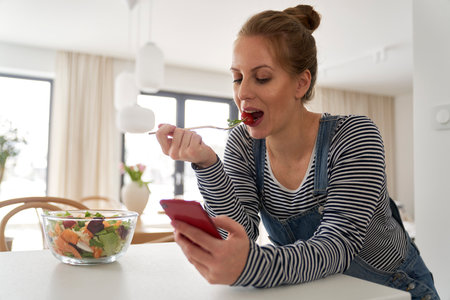 Caucasian woman in the kitchen eating a salad and browsing phoneの写真素材