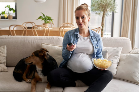 Pregnant woman watching TV and eating crisps with a dogの写真素材